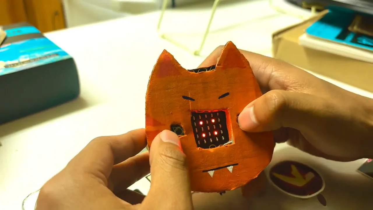A hand holding a small cardboard cat enclosure with a micro:bit LED face showing through the front